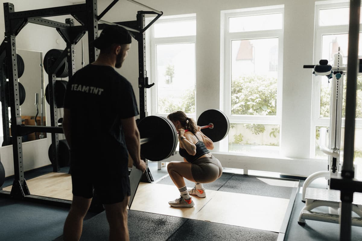 Ein Trainer aus dem Team TNT begleitet eine Sportlerin bei der Kniebeuge mit der Langhantel